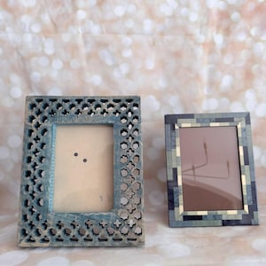 May include: Two decorative picture frames. One frame is a distressed blue with an intricate floral cutout design. The second frame has a mosaic pattern in shades of blue, gray, and beige. Both frames are rectangular and designed to hold photos.