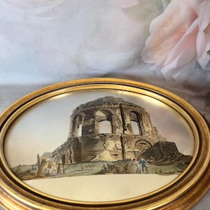May include: An oval-shaped wall plaque featuring a detailed print of ancient stone ruins with arched openings. The scene includes small figures standing near the structure. The print is encased in a polished gold-toned frame with a smooth finish.