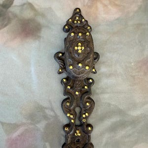 May include: A dark brown, cast metal door plate and knob assembly featuring an ornate, scrollwork design. The surface is embellished with numerous small, bright yellow, circular accents embedded throughout the textured, dark metal frame.
