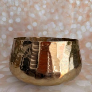 May include: A hammered gold-colored bowl with a wide, rounded shape. The bowl has a textured surface and reflects light, creating highlights and shadows. The bowl's design is simple yet elegant, suitable for various decorative purposes.