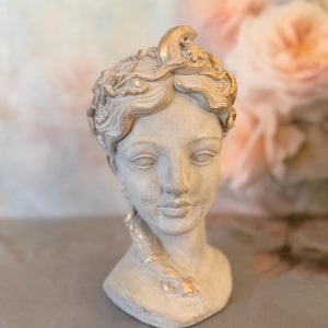 May include: A light gray bust of a person with a serene expression, featuring a braided hairstyle adorned with gold-painted accents, including a crescent moon and star motif integrated into the hair, with a smooth, matte finish on the facial features.