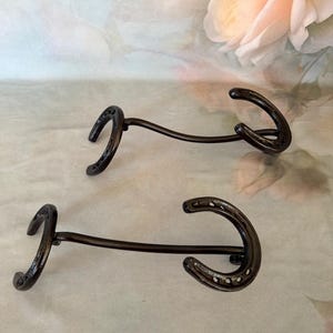 May include: Two black metal wall hooks, each constructed from a curved horseshoe attached to a straight metal rod that connects to a second, smaller horseshoe-shaped hook at the opposite end, featuring a dark, smooth, uniform finish throughout.