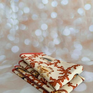 May include: A stack of four folded cream-colored napkins with a coral and seashell pattern. The napkins have a rust-colored coral design and a red border. The background is a soft, blurred bokeh effect.