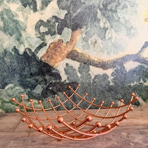 May include: A copper-colored metal fruit bowl with a lattice design. The bowl has a curved shape with small spheres at the intersections of the metal bars. The background features a textured wall with a watercolor-style design.