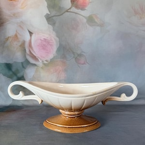 May include: A ceramic gravy boat or serving dish featuring a cream-colored interior and exterior with a fluted design. The base and lower portion are finished in a speckled brown glaze. It includes two curved handles on opposite sides.