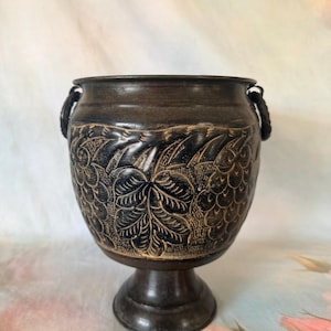 May include: A dark brown, footed metal vessel featuring a wide, rounded body with a flared rim and two small side handles. The exterior surface is decorated with an intricate, carved-style pattern of textured leaves and scale-like motifs.