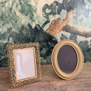 May include: Two gold-colored picture frames on a wooden surface. One frame is rectangular with an ornate, scrolled design. The other is a gold-colored, oval frame with a ridged border. The background features a green and brown abstract painting.