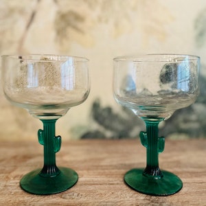 May include: Two clear glass margarita glasses with green cactus-shaped stems and bases. The glasses are empty and sitting on a wooden surface.