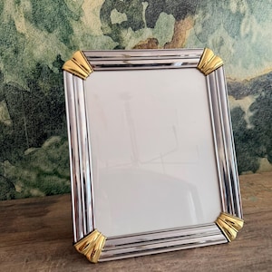 May include: A rectangular picture frame featuring a polished silver-tone metallic finish with vertical ribbed detailing. The frame is accented by four gold-tone fan-shaped corner pieces that provide a distinct contrast to the silver frame body.