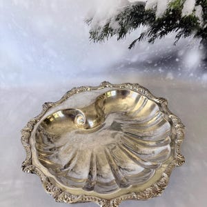 May include: A silver-toned metal serving tray shaped like a large seashell. The tray features a scalloped edge with intricate embossed floral detailing along the rim and a smooth, curved interior surface that mimics the natural ridges of a shell.