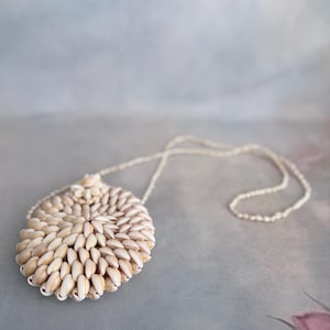 May include: A handcrafted necklace featuring a large, oval pendant made of small, cream-colored seashells. The pendant is attached to a delicate, beaded chain. The background is a soft, blurred mix of gray and blue.