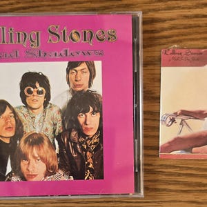 May include: A CD case with the text "Rolling Stones Mad Shadows" and a photo of the band. Next to it is a small album cover with a woman in a swimsuit and headphones.