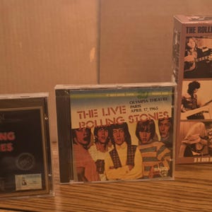 Könnte beinhalten: Drei Musikveröffentlichungen der Rolling Stones. Die erste ist eine CD-Hülle mit den Worten "Rolling Stones" auf schwarzem Hintergrund. Die zweite ist eine CD-Hülle mit den Worten "The Live Rolling Stones" und einem Foto der Band. Die dritte ist eine DVD-Box.