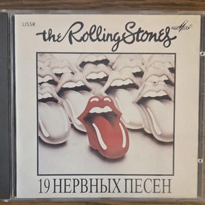May include: A CD case featuring The Rolling Stones album cover with the text USSR, the Rolling Stones logo, and 19 НЕРВНЫХ ПЕСЕН. The artwork displays a central red tongue and lips graphic surrounded by multiple grayscale versions of the same icon.