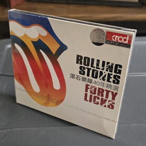 Puede incluir: Una caja de CD del álbum "Forty Licks" de The Rolling Stones. La portada presenta el icónico logotipo de la lengua y los labios de la banda en rojo, naranja y amarillo sobre un fondo blanco. El título del álbum y el nombre de la banda están impresos en negro.