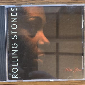 May include: A compact disc case featuring the Rolling Stones album "Miss You." The cover art displays a side profile of a person's face in warm tones. The band's name is vertically printed in white, with the song title in cursive.