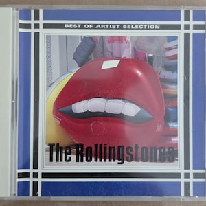 Puede incluir: Una caja de CD con una boca roja de gran tamaño y dientes blancos. El texto "The Rollingstones" está debajo. La caja tiene un borde azul con detalles en blanco y negro, y el texto "BEST OF ARTIST SELECTION" en la parte superior.