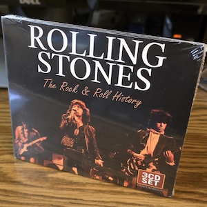 May include: A sealed CD set of The Rolling Stones' "The Rock & Roll History." The album cover is black with white text that reads "Rolling Stones" and "The Rock & Roll History." The cover features a live performance photo of the band. The set includes 3 CDs.