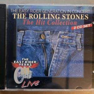 Puede incluir: Una caja de CD de "The Rolling Stones: The Hit Collection." La portada muestra un primer plano de vaqueros azules. El texto dice "The Easy Rider Generation in Concert" y "2 CD SET." El CD está etiquetado como "Live."