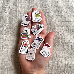 May include: A collection of white ghost stickers with black eyes and various accessories. Some ghosts hold books, a witch hat, a baseball cap, and a wand. The stickers are designed with a cartoon style and are suitable for Halloween or spooky-themed crafts.