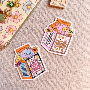 Puede incluir: Dos pegatinas coloridas con forma de cajas de zumo. Una dice "Self Love Juice" y la otra "Happy Juice". Ambas tienen un diseño retro con estrellas, arcoíris y las palabras "Drink Up!"