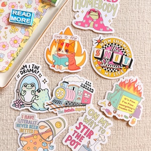 Puede incluir: Una colección de pegatinas coloridas con varios diseños y texto. Las pegatinas incluyen una rana en una manta, un gato leyendo en llamas, un tren, una rana en un espejo, un cubo de basura en llamas y textos como "Certified Homebody" y "Read More".