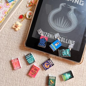 Puede incluir: Un lector de libros electrónicos con una funda dorada está rodeado de dijes de libros en miniatura y un dije decorativo. El lector muestra el texto "Sunrise on the Reaping" de Suzanne Collins. Los dijes presentan portadas de libros con varios títulos.
