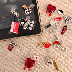 May include: A collection of gothic-themed charms and accessories, including a red heart, a knife with blood splatter, a skull, a butterfly, and a coffin. Some charms are attached to clear clips. The word "LOVEBOOKS" is on a gold bar.