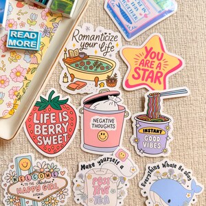 Puede incluir: Una colección de pegatinas coloridas con varios diseños y texto. Las pegatinas incluyen una fresa con "Life is Berry Sweet", una estrella con "You Are A Star" y una ballena con "Everything Whale Be Okay". También incluye libros en miniatura.