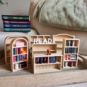 Puede incluir: Una colección de estanterías de madera en miniatura, algunas llenas de libros pequeños. La palabra "READ" está escrita con letras blancas. Se ve una pila de libros con títulos como "Heir of Fire" y "Throne of Glass". También hay un pequeño oso de peluche y una figura de perro rosa.