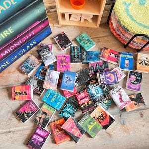 May include: A collection of miniature books with colorful covers, scattered on a wooden surface. Titles include "Harry Potter," "A Court of Thorns and Roses," and "Housemaid." A small wooden bookshelf and a stack of smiley face coasters are also visible.