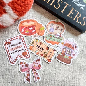 May include: A collection of fall-themed stickers featuring pumpkins, a bow, and a snow globe. The stickers include text such as "Cozy Book Club" and "I stopped my cozy read here." The color palette includes orange, pink, and brown.