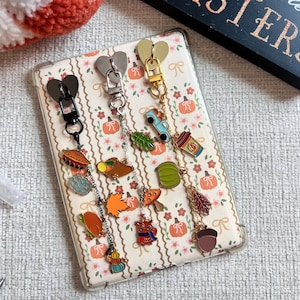 Puede incluir: Una colección de llaveros con dijes temáticos de otoño, que incluyen calabazas, hojas y bellotas. Los llaveros tienen cierres en forma de corazón en negro, plateado y dorado. El fondo es una funda de teléfono con un estampado de calabazas y flores.