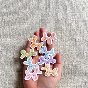 Puede incluir: Una mano sosteniendo una colección de pegatinas de animales de globo de colores. Las pegatinas tienen forma de perro y son de varios tonos de rosa, azul, amarillo y naranja.