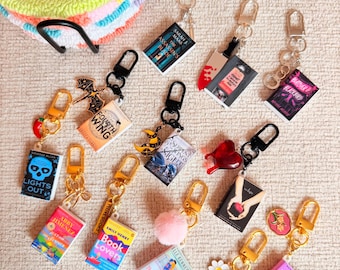 May include: A collection of miniature book keychains featuring various titles like The Assassin's Blade, Fourth Wing, and Book Lovers. Each keychain includes a metal clasp and unique charms such as a knife, heart, pom-pom, or flower, attached to a small rectangular book replica.