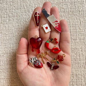 May include: A collection of red and silver charms, including a coffin, a bloody knife, a heart, a playing card, a chili pepper, lips, a rose, a bow, and a butterfly. The charms are small and detailed, with gold accents.