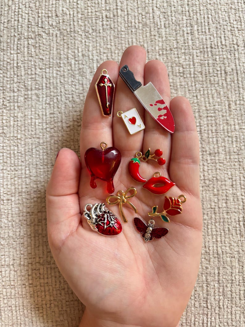 May include: A collection of red and silver charms, including a coffin, a bloody knife, a heart, a playing card, a chili pepper, lips, a rose, a bow, and a butterfly. The charms are small and detailed, with gold accents.