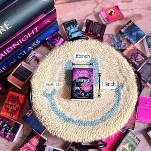 May include: A miniature book titled "Empire of Storms" by Sarah J. Maas, measuring .85 inches wide and 1.3 inches tall, rests on a yellow rug with a blue smiley face design. Surrounding the rug are several other miniature books.