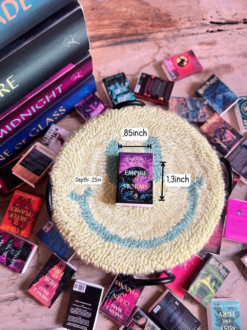 May include: A miniature book titled "Empire of Storms" by Sarah J. Maas, measuring .85 inches wide and 1.3 inches tall, rests on a yellow rug with a blue smiley face design. Surrounding the rug are several other miniature books.