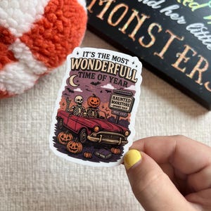 以下が含まれることがあります： 「It's the Most Wonderful Time of Year」のテキストが入ったハロウィーンをテーマにしたステッカー。骸骨とジャック・オー・ランタンが運転する赤い車が描かれています。カボチャと、お化け屋敷の書店を示す看板も含まれています。
