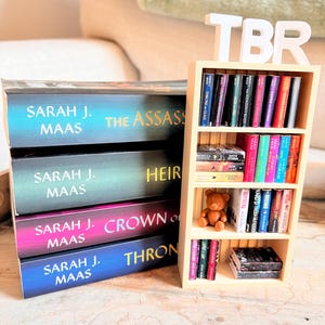 May include: A stack of books by Sarah J. Maas, with titles including "The Assassin's Blade," "Heir of Fire," and "Crown of Midnight." A small wooden bookshelf displays more books and a teddy bear. A white "TBR" sign sits atop the bookshelf.