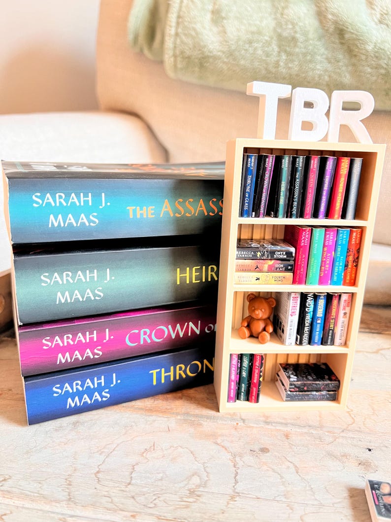 May include: A stack of books by Sarah J. Maas, with titles including "The Assassin's Blade," "Heir of Fire," and "Crown of Midnight." A small wooden bookshelf displays more books and a teddy bear. A white "TBR" sign sits atop the bookshelf.
