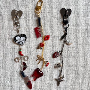 May include: Three keychains with heart-shaped clasps. One features a black heart with skeletons, a chili pepper, and a knife. Another has a bloody knife, red lips, and a heart. The third has a crow, skull, and star.