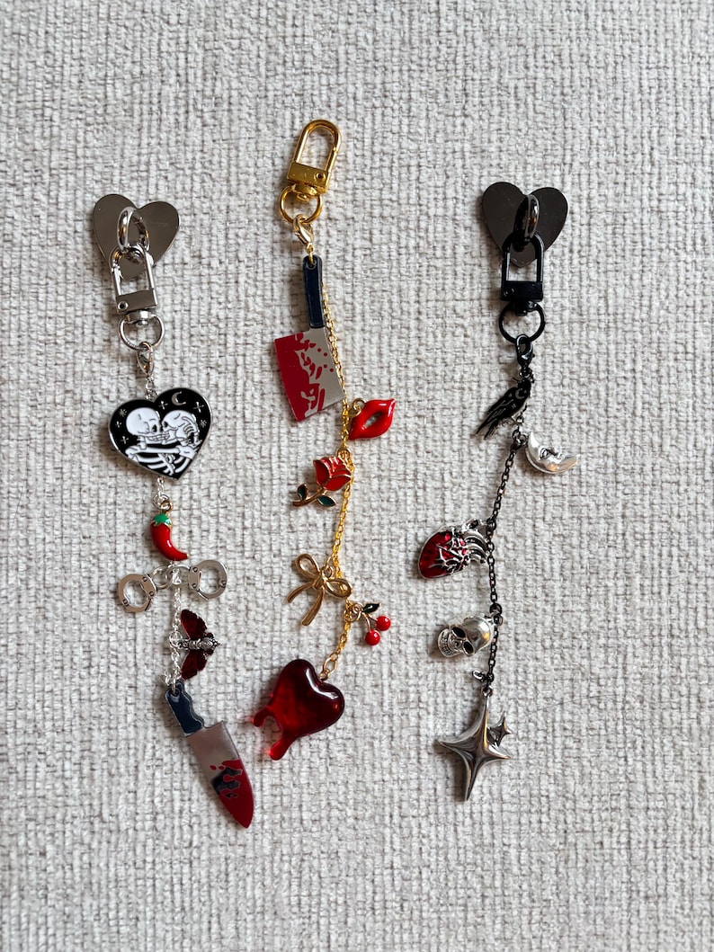 May include: Three keychains with heart-shaped clasps. One features a black heart with skeletons, a chili pepper, and a knife. Another has a bloody knife, red lips, and a heart. The third has a crow, skull, and star.