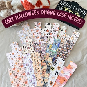 May include: A collection of Halloween-themed phone case inserts with various patterns, including pumpkins, ghosts, and witches. The inserts are displayed with two decorative plush pumpkins and a sign that reads "Here Lives the Wicked Witch."