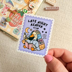 May include: A sticker with a purple border featuring a cartoon duck reading a book under a crescent moon and stars. The text reads "LATE NIGHT READER CLUB" and "SLEEP IS OPTIONAL. BOOKS ARE NOT." Also visible are a phone case with stickers and a keychain.