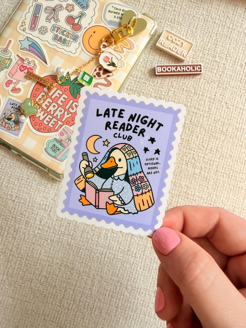 May include: A sticker with a purple border featuring a cartoon duck reading a book under a crescent moon and stars. The text reads "LATE NIGHT READER CLUB" and "SLEEP IS OPTIONAL. BOOKS ARE NOT." Also visible are a phone case with stickers and a keychain.