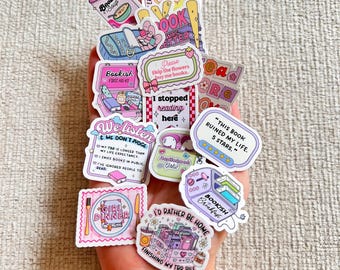 Op de afbeelding: Een verzameling kleurrijke stickers met verschillende thema's en zinnen over boeken. De stickers bevatten tekst als "Bookish Soup", "Certified Bookworm" en "I'd Rather Be Home Finishing My TBR Pile". De stickers worden in een hand gehouden.