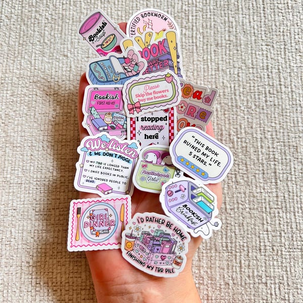 Peut inclure: Un ensemble d'autocollants colorés sur le thème des livres, avec des phrases telles que "Bookish Soup", "Certified Bookworm" et "I'd Rather Be Home Finishing My TBR Pile". Les autocollants sont tenus dans une main.