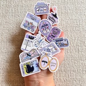 Puede incluir: Una colección de pegatinas con temática de fantasía en varias formas y tamaños. Los diseños incluyen un caldero, un libro, una lata de refresco y una carta del tarot. Las pegatinas presentan colores morado, blanco y negro, con texto como "Fantasy Bookpol" y "Fantasy girlie."